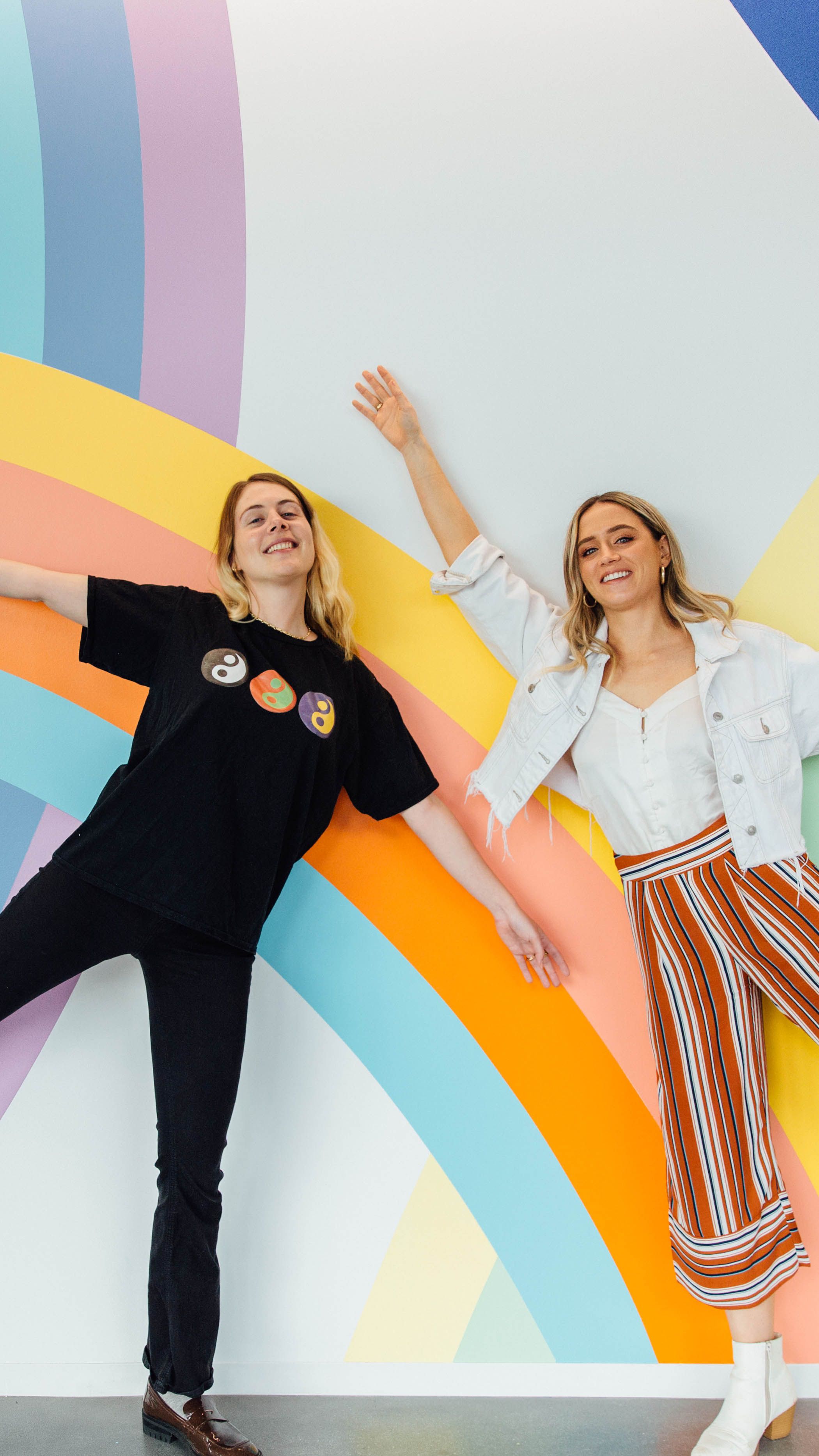 Two young female employees pose in front of a rainbow wall